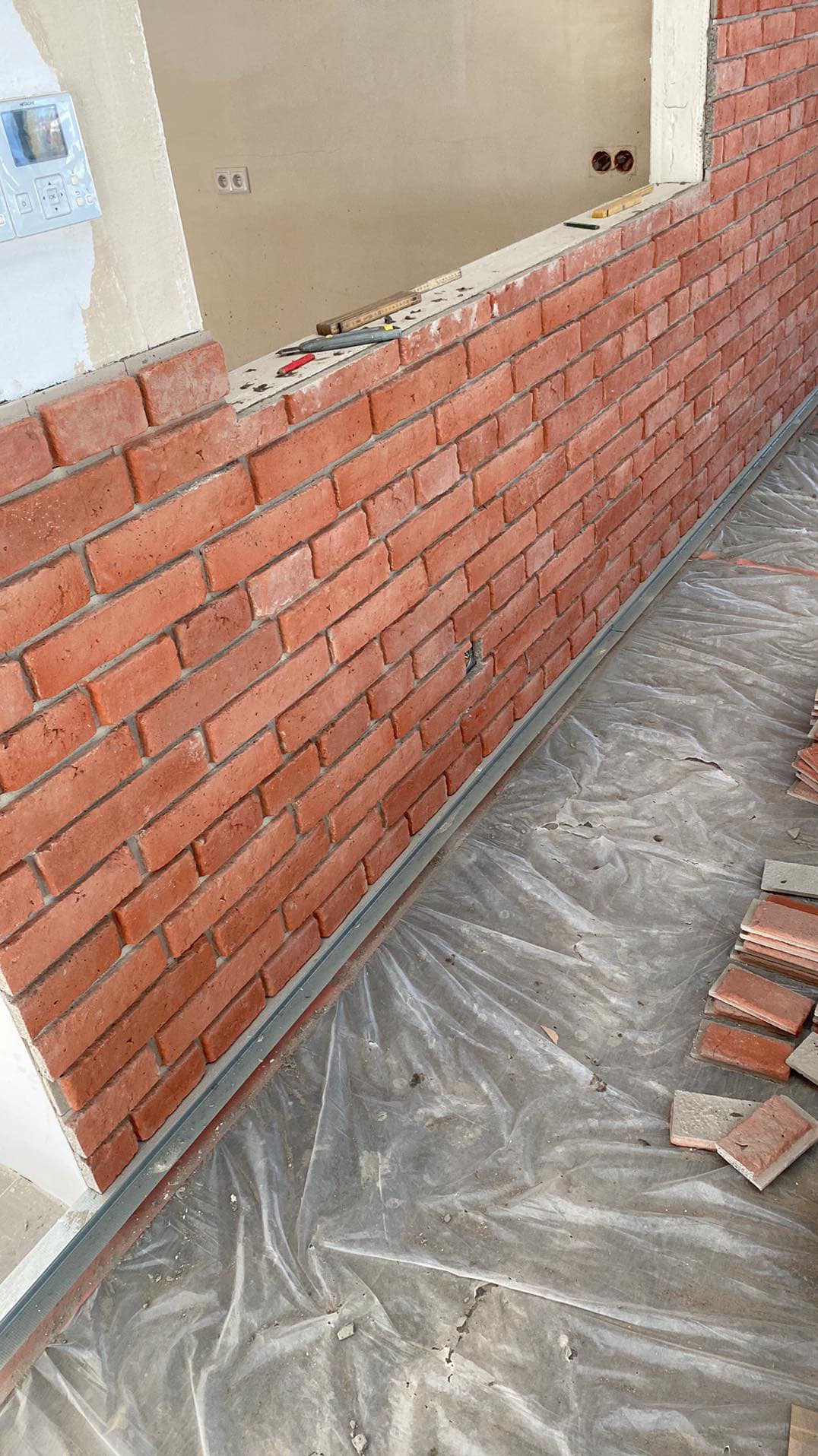 Montaż okładziny z cegły klinkierowej na ścianie wewnętrznej. Widoczne narzędzia i fragmenty płytek. Prace w toku, podłoga zabezpieczona folią.