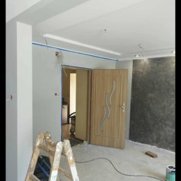 Daniel Kidziak - Wnętrze w trakcie malowania: drabina, ściany z taśmą malarską, częściowo pomalowana ściana dekoracyjna, otwarte drzwi. Widoczne kable i puszki elektryczne.