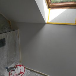 Daniel Kidziak - Ściana po malowaniu na biało, zabezpieczona folią i taśmą malarską, widoczne narzędzia malarskie i okno dachowe z widokiem na zieleń.