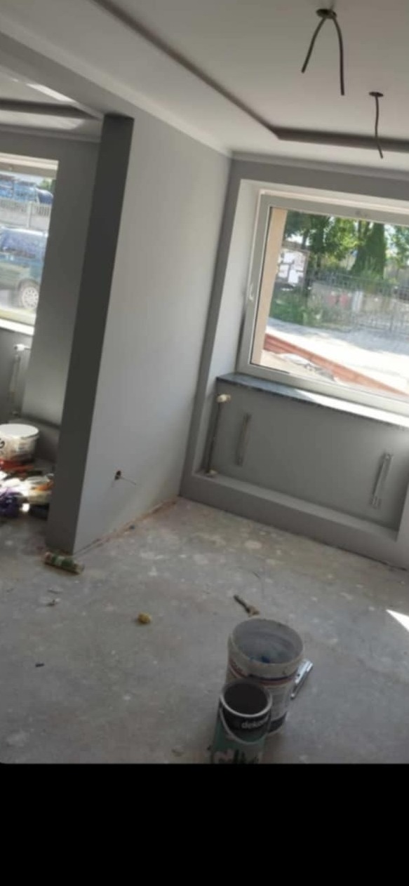 Szare, świeżo pomalowane wnętrze pokoju z oknami, widoczne puszki z farbą na podłodze. Minimalistyczny wystrój sufitu.