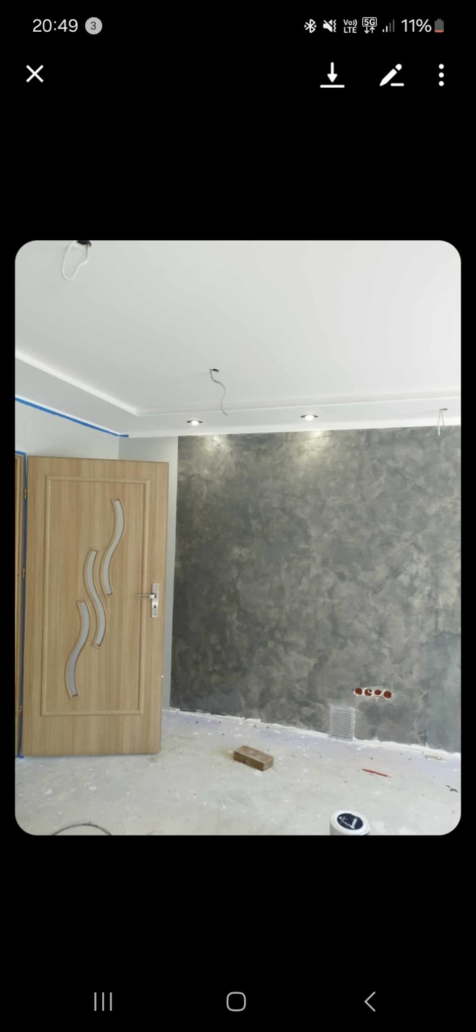 Wykończone wnętrze pokoju z dekoracyjną szarą ścianą, drewnianymi drzwiami z przeszkleniami i listwami sufitowymi. Widoczne gniazdka elektryczne i fragment podłogi w trakcie prac.