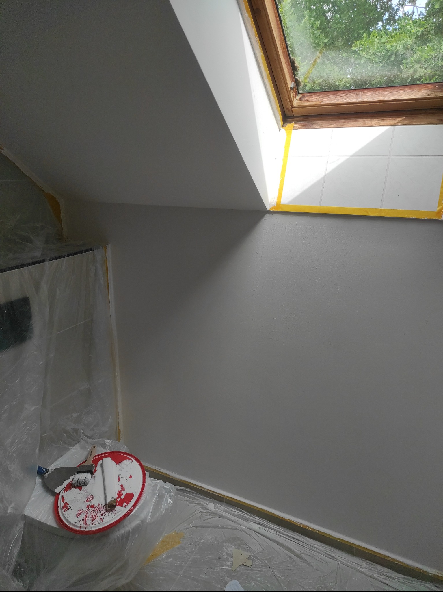 Ściana po malowaniu na biało, zabezpieczona folią i taśmą malarską, widoczne narzędzia malarskie i okno dachowe z widokiem na zieleń.