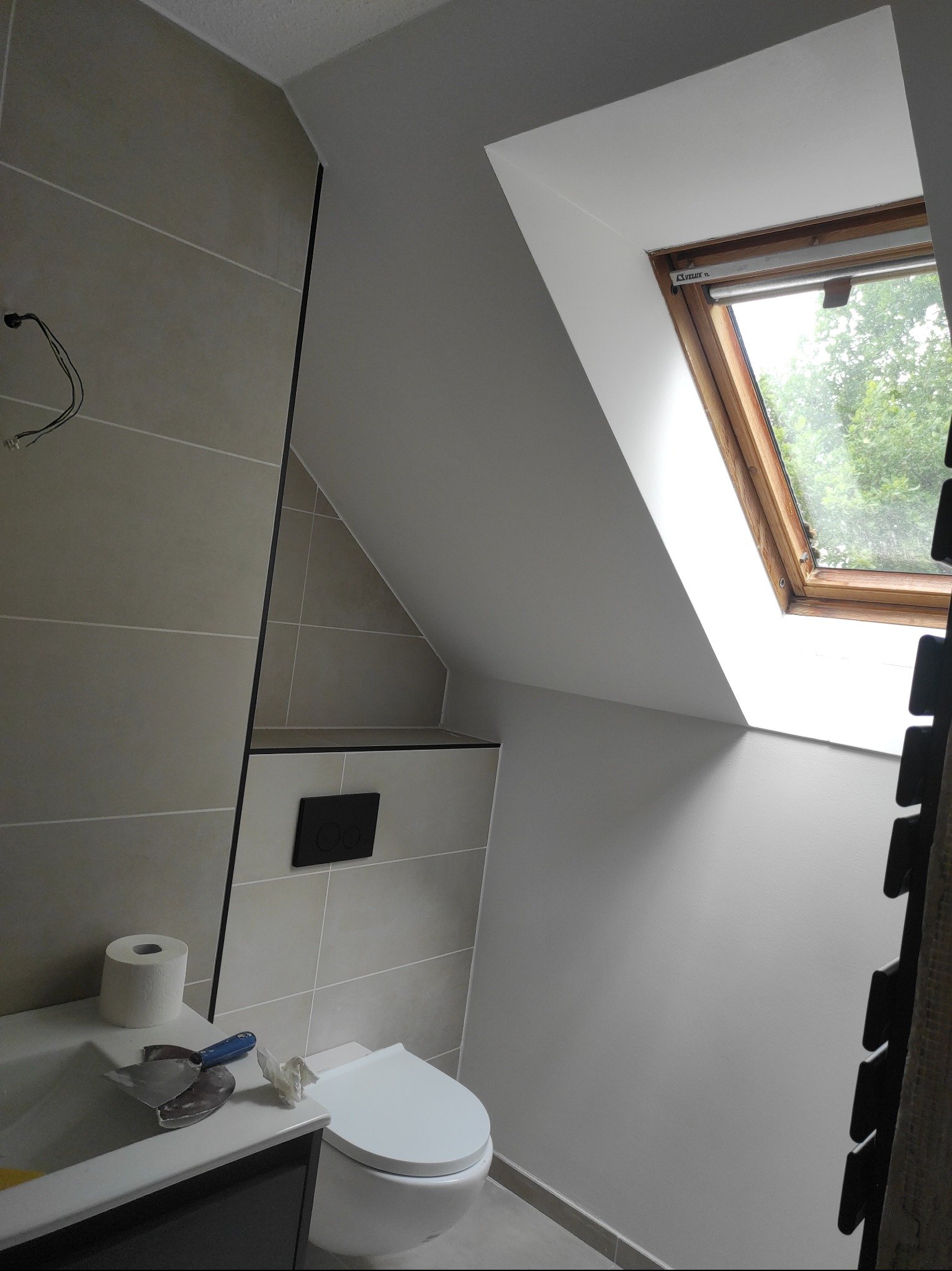 Wykończona łazienka na poddaszu z oknem dachowym. Widoczne płytki, toaleta i narzędzia na blacie. Świeżo pomalowane ściany i wykończone detale.