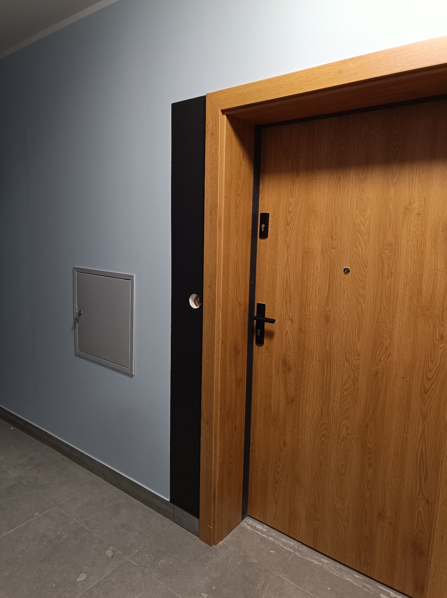 Zamontowane drzwi wejściowe z drewnianą okleiną, czarną ościeżnicą i szarą ścianą. Widoczny fragment podłogi z szarych płytek. Na ścianie rewizja.