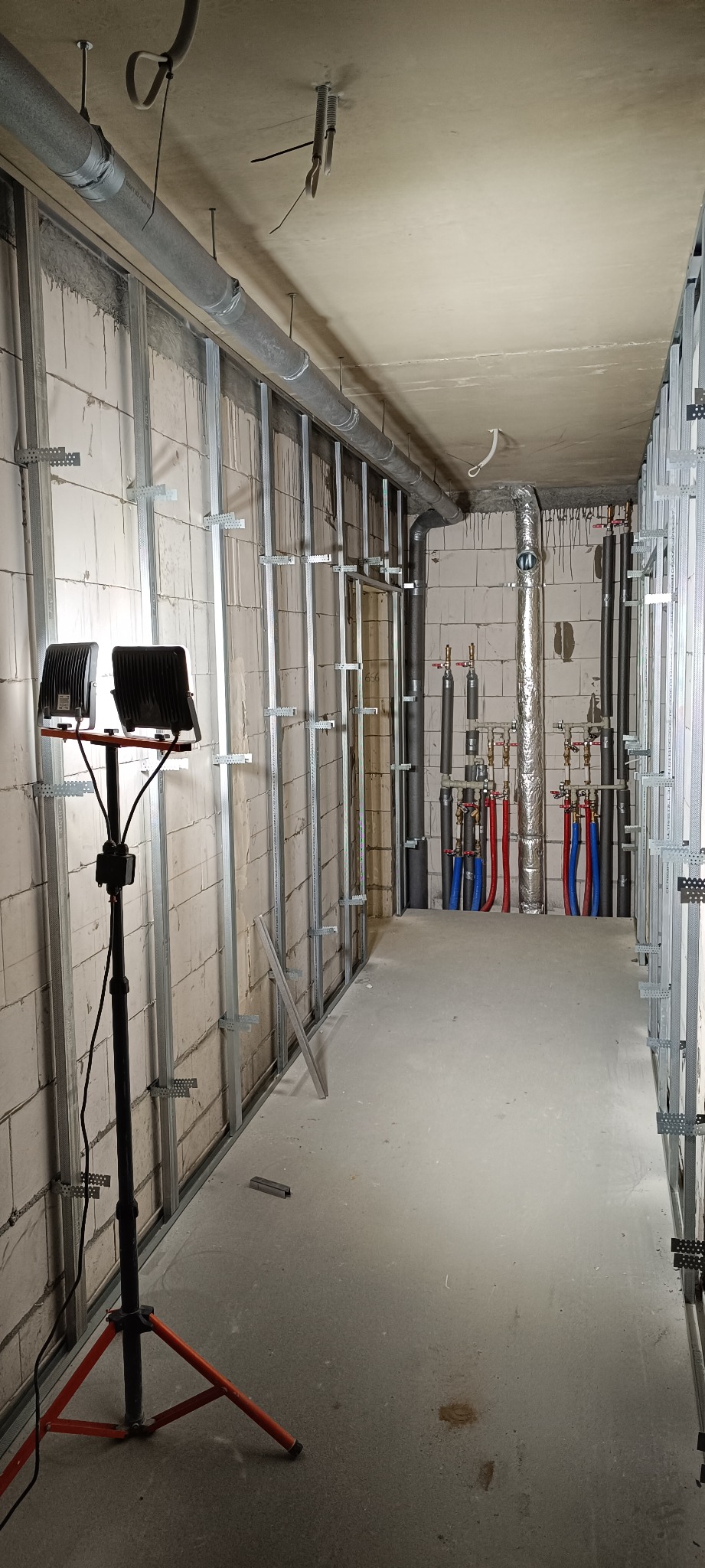 Wnętrze w trakcie prac: metalowy stelaż pod płyty G-K na ścianach z białego bloczka, instalacja wodna z rurami w kolorach niebieskim i czerwonym, oświetlenie budowlane.