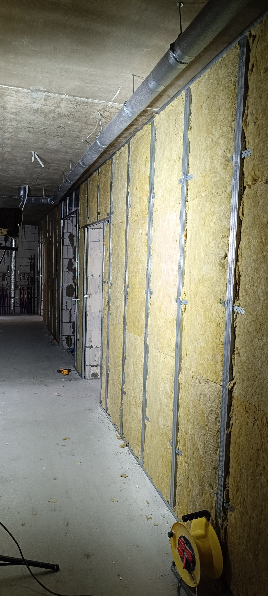 Montaż izolacji z wełny mineralnej na metalowym stelażu w surowym wnętrzu. Widoczne narzędzia i instalacje pod sufitem. Oświetlenie surowe, budowlane.