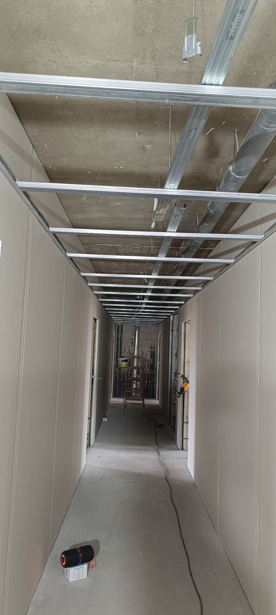 Korytarz w trakcie prac wykończeniowych. Widoczne ściany z płyt gipsowo-kartonowych, metalowa konstrukcja sufitu podwieszanego i elementy instalacji. W tle drabina i narzędzia.