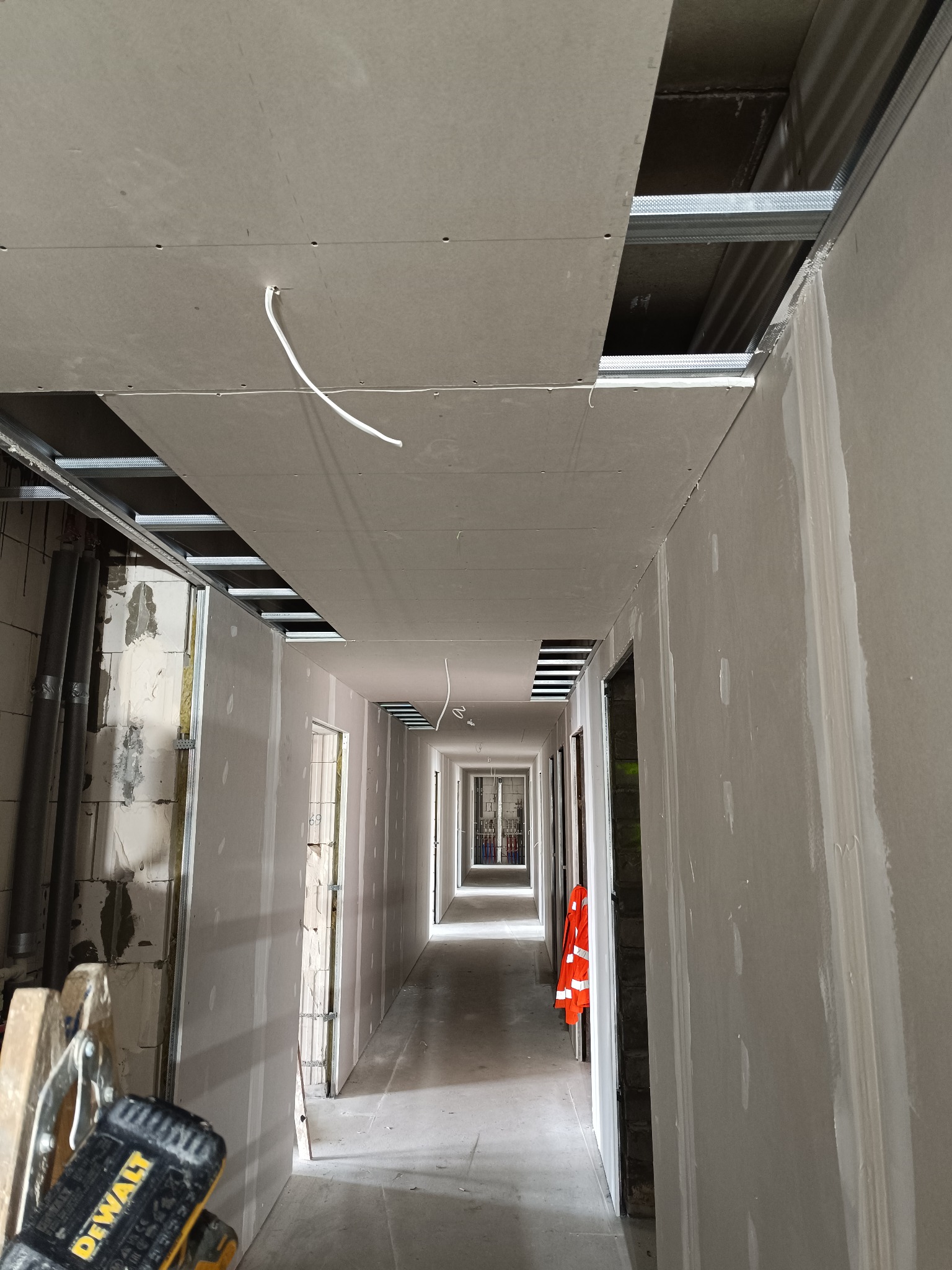Korytarz w trakcie montażu płyt gipsowo-kartonowych na ścianach i suficie, widoczne profile metalowe i przewody elektryczne. W tle perspektywa z otwartymi drzwiami.