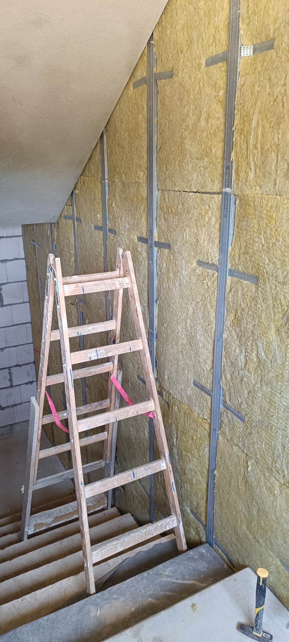 Ocieplenie poddasza wełną mineralną, widoczne metalowe profile i drabina. Prace wykończeniowe na poddaszu, izolacja termiczna ścian i stropu.