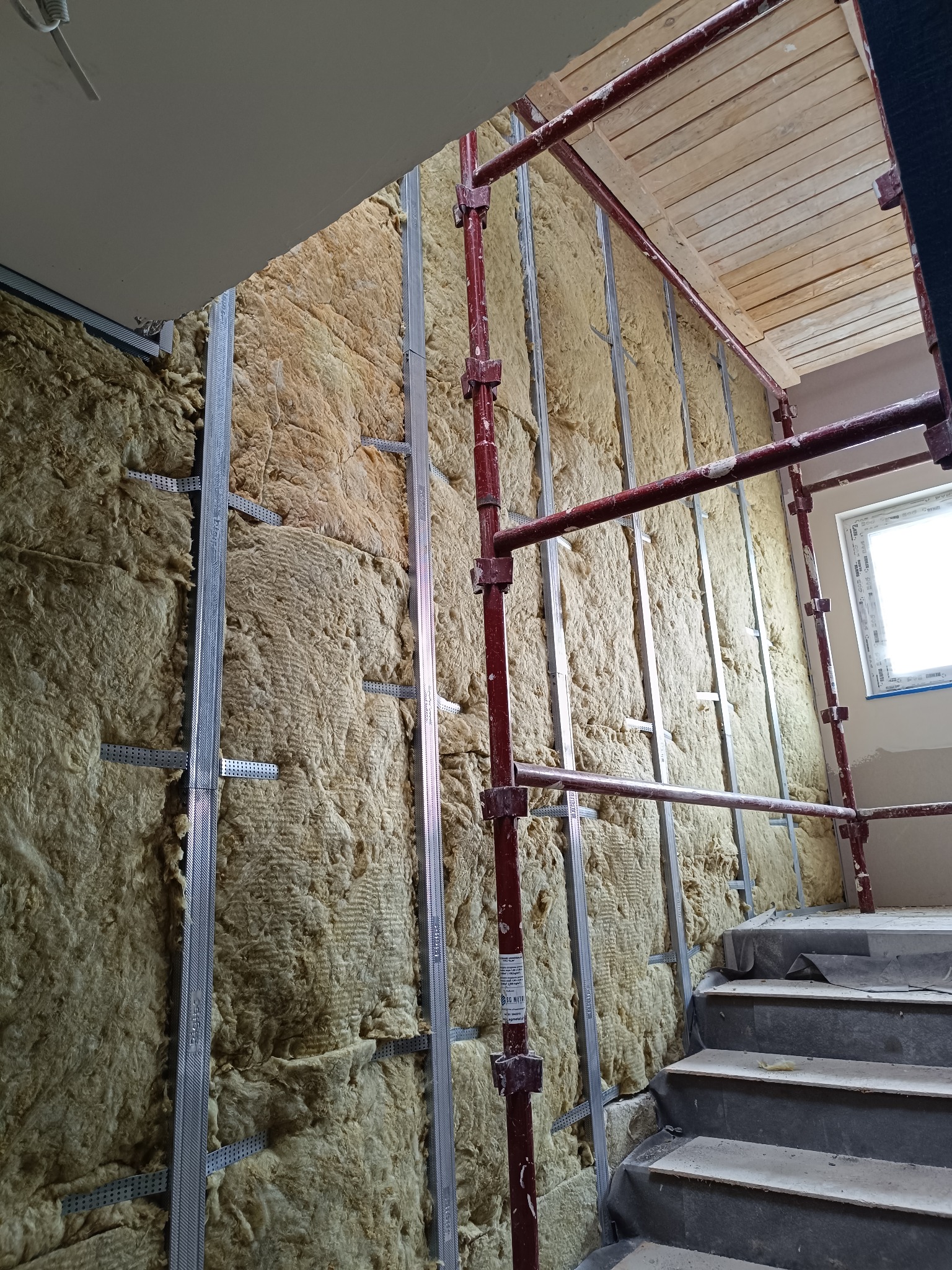 Ocieplenie ścian wełną mineralną na metalowym stelażu, widoczne rusztowanie i schody. Prace wykończeniowe w klatce schodowej budynku.