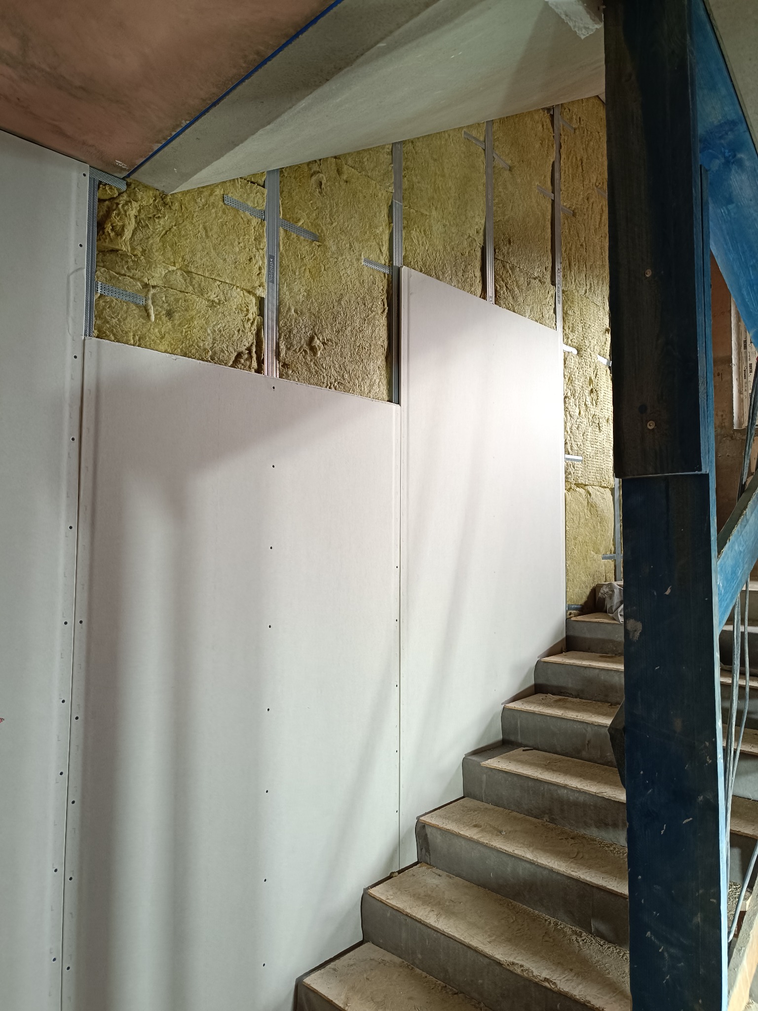 Montaż płyty karton gips na ścianie z izolacją, widoczne schody. Metalowe profile, surowy stan wykończenia. Ujęcie z dołu, perspektywa schodów.