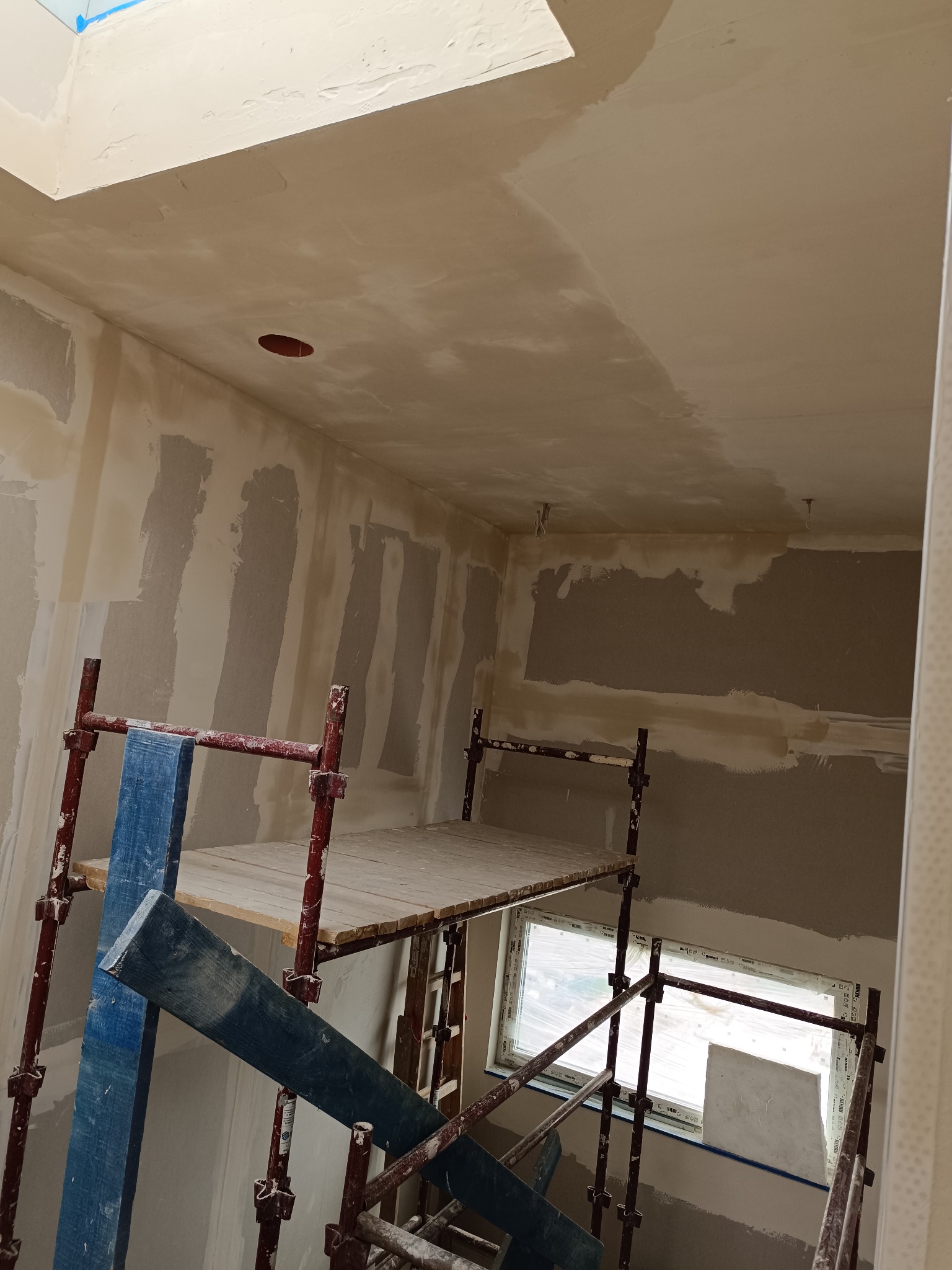 Wnętrze w trakcie szpachlowania: ściany i sufit pokryte gładzią, widoczne rusztowanie z drewnianym podestem, okno zabezpieczone folią. Otwór w suficie pod instalację.