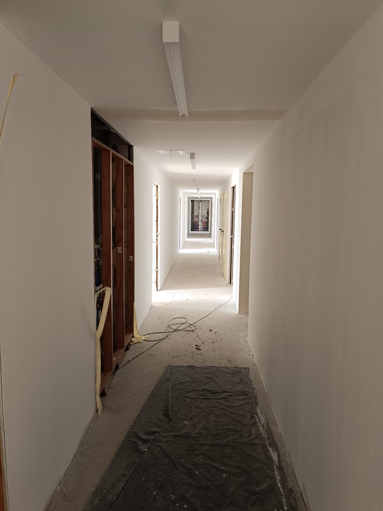 Korytarz w trakcie remontu: świeżo pomalowane ściany na biało, widoczne instalacje w ścianie, zabezpieczona podłoga folią. Perspektywa długa, oświetlenie sufitowe.