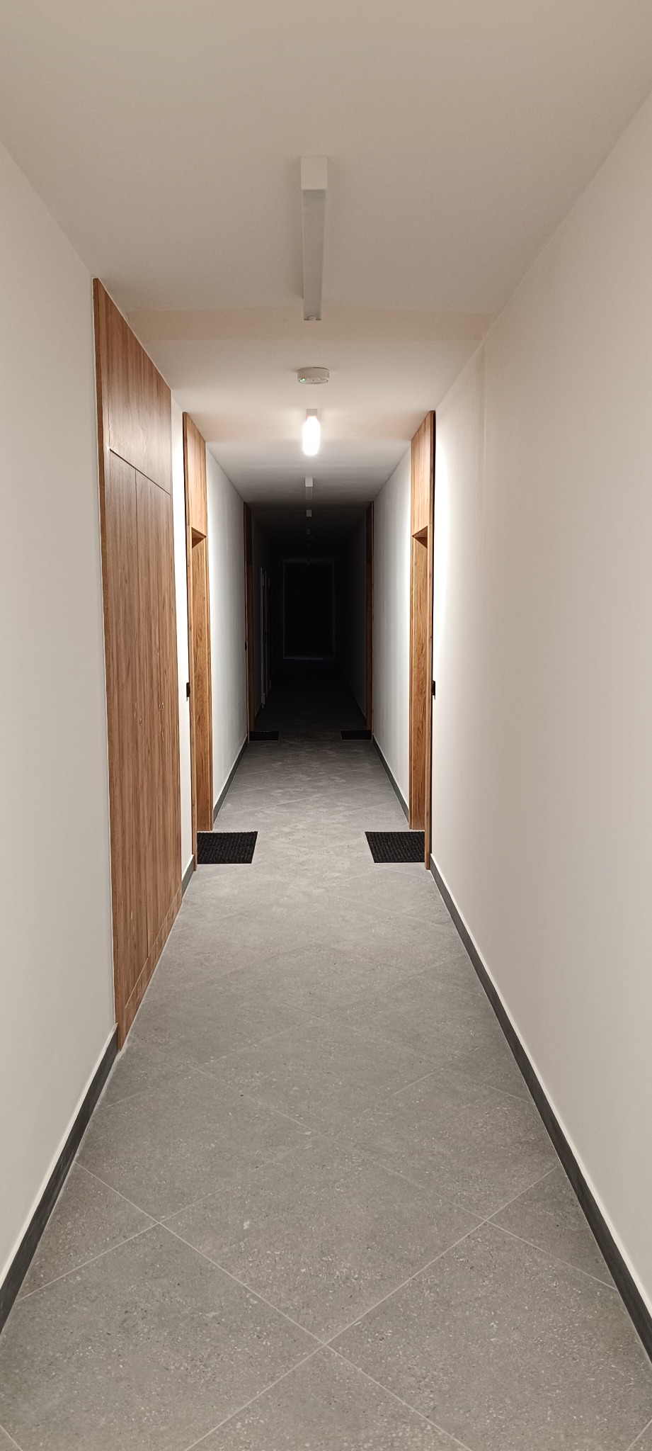 Długi korytarz z szarą podłogą w romby, białymi ścianami i drewnianymi drzwiami po bokach. Widoczne lampy sufitowe i wycieraczki przed drzwiami. Perspektywa zbieżna w oddali.