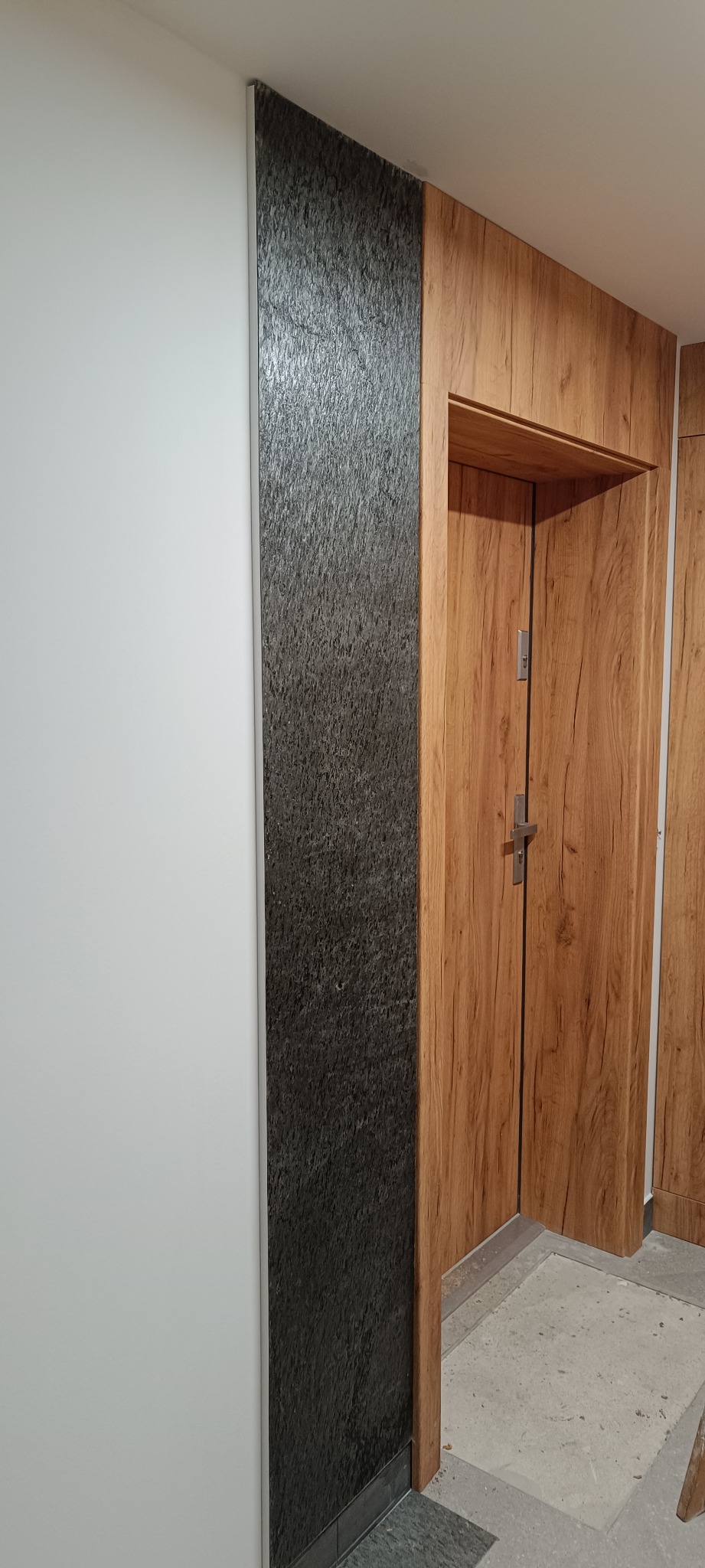 Wykończone wnętrze: drewnopodobne obramowanie drzwi, ściana z czarnej, strukturalnej okładziny i gładka, biała ściana. Kontrast faktur i kolorów.