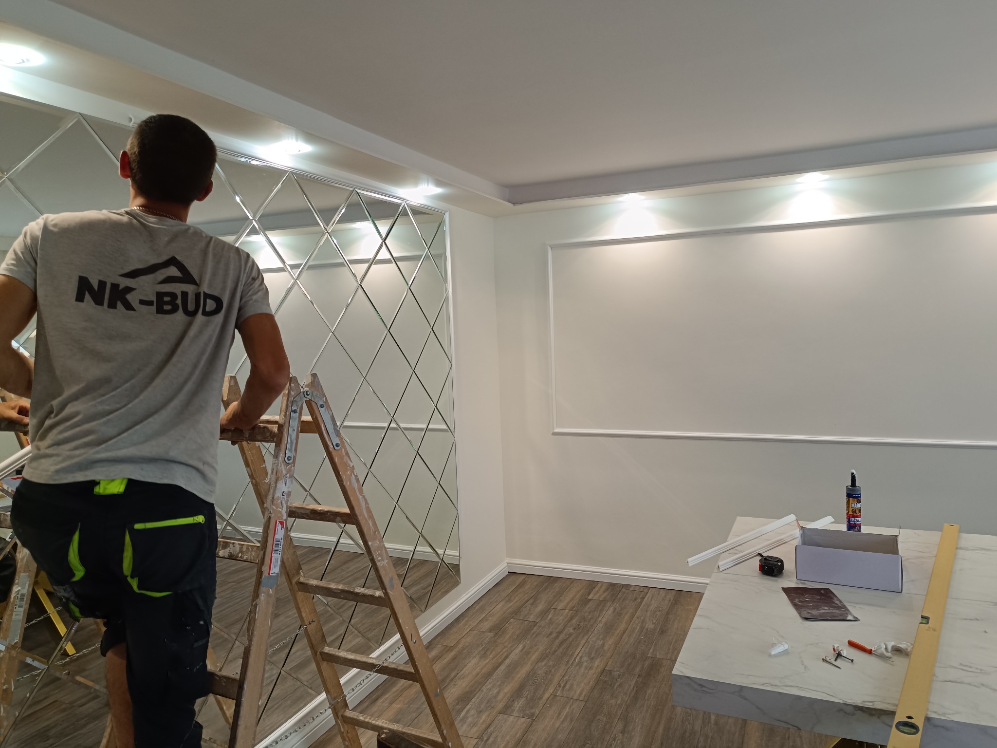 Montaż dekoracyjnych luster na ścianie w romby. Widoczny pracownik na drabinie, narzędzia i materiały przygotowane do montażu. Nowoczesne wykończenie wnętrza.