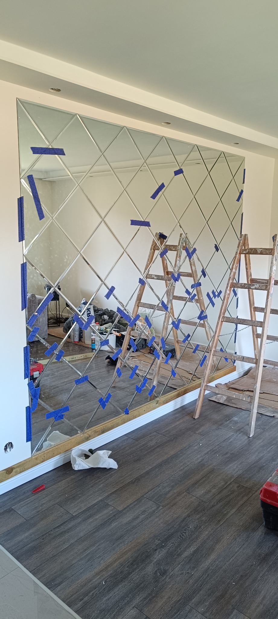 Montaż dekoracyjnego lustra z rombów na ścianie, zabezpieczonego niebieską taśmą. Dwie drewniane drabiny opierają się o lustro. Widoczne narzędzia i podłoga imitująca drewno.