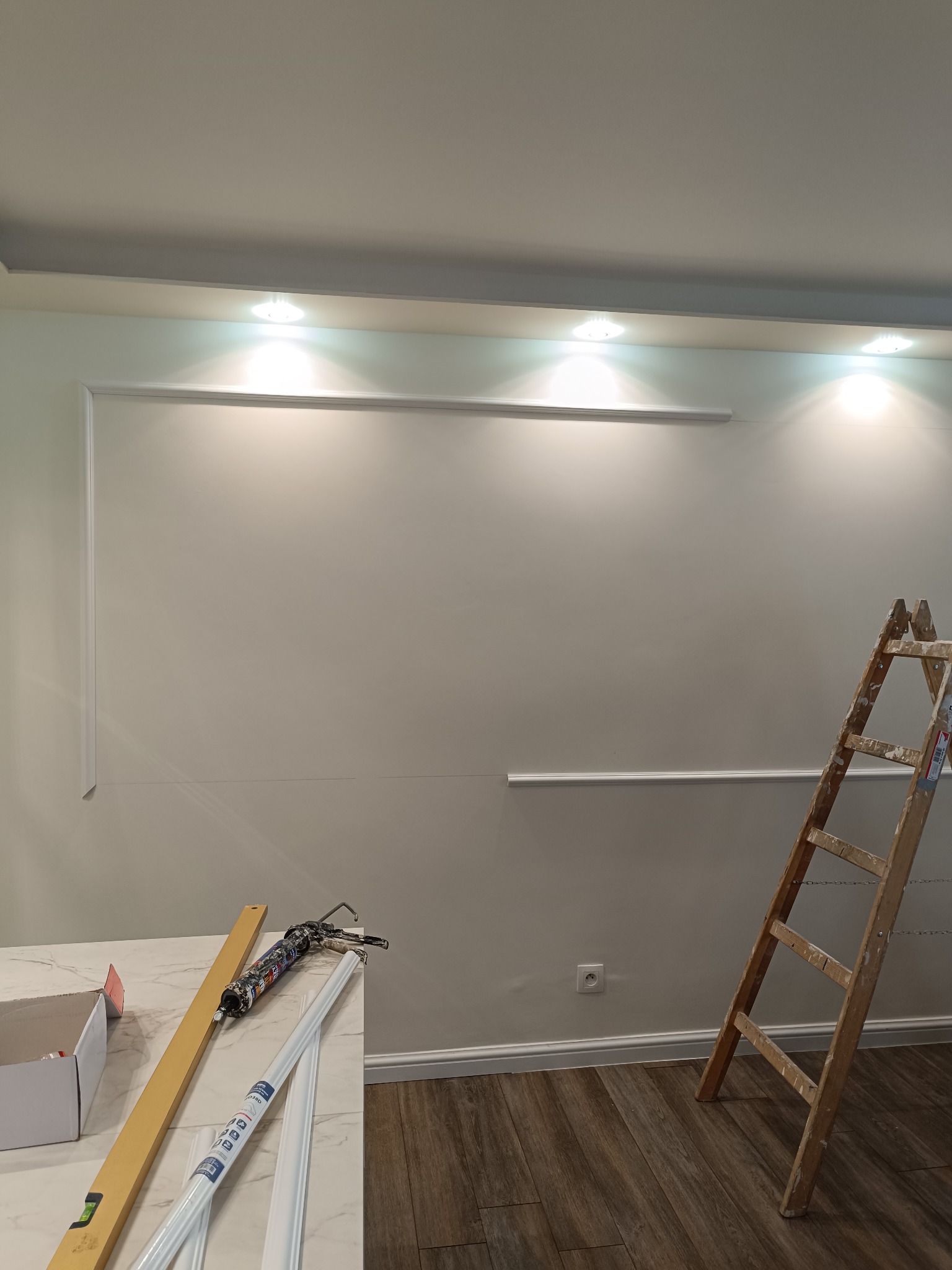 Montaż sztukaterii ściennej w jasnym wnętrzu. Widoczna drabina, listwy przypodłogowe i narzędzia na blacie. Częściowo zamontowane listwy dekoracyjne.