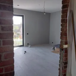 Szare ściany w pokoju po malowaniu, widok przez ceglany otwór drzwiowy. Na podłodze panele, widoczne narzędzia i puszka farby. W tle okno z widokiem na zieleń.