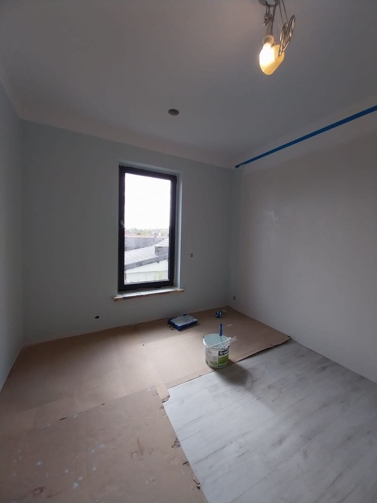 Pokój po malowaniu ścian na biało, z zabezpieczoną podłogą, widoczne narzędzia malarskie i taśma malarska przy suficie. Świeże wykończenie wnętrza.