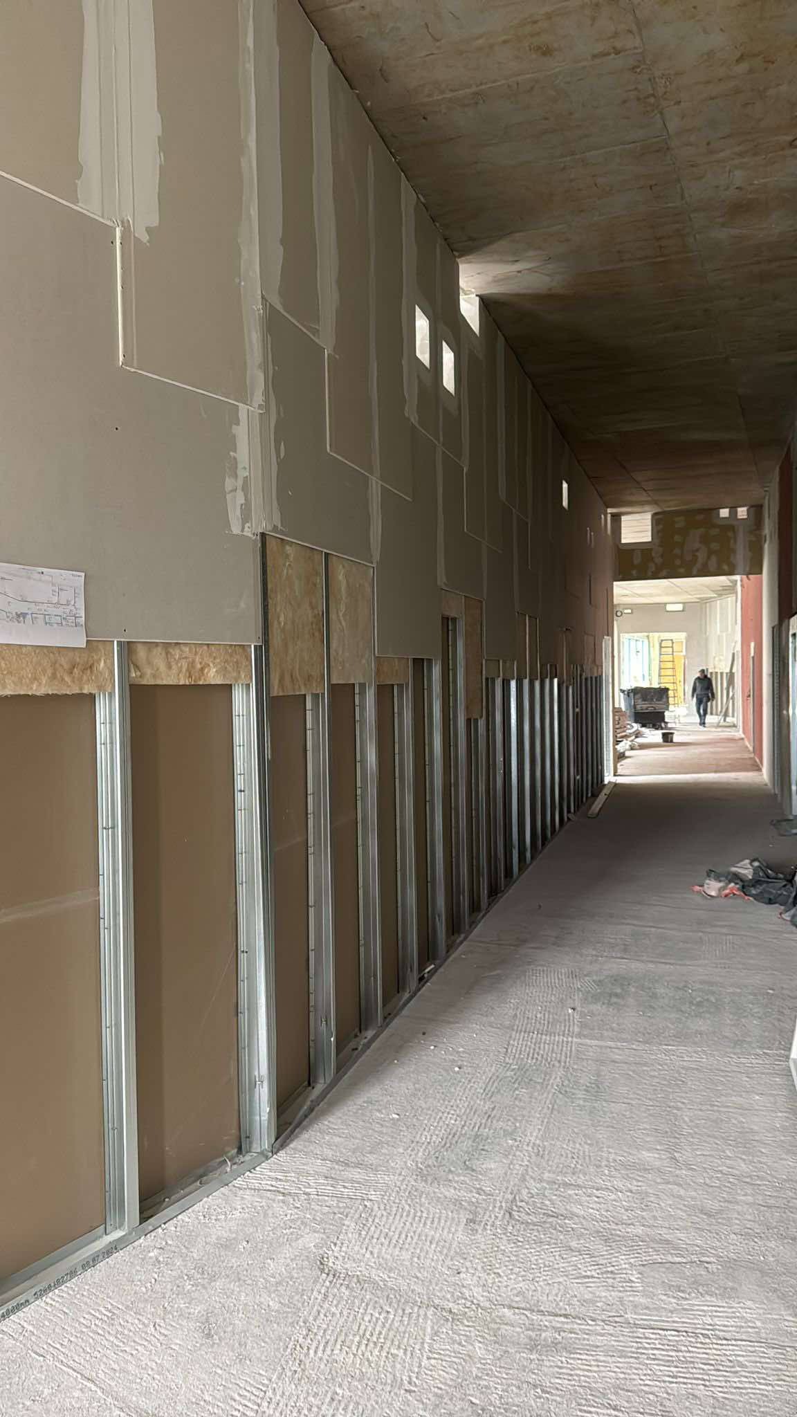 Montaż płyt gipsowo-kartonowych w korytarzu, widoczne profile i izolacja. Prace w toku, surowy betonowy sufit, w tle osoba i wózek budowlany.