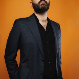 Alicja Kopiec - Mężczyzna z brodą w ciemnym garniturze i czarnej koszuli, patrzy w górę na pomarańczowym tle.