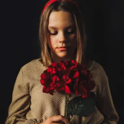 Portret młodej kobiety z zamkniętymi oczami, trzymającej czerwoną hortensję na ciemnym tle, z delikatnym oświetleniem podkreślającym rysy twarzy i fakturę sukienki.