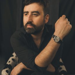 Alicja Kopiec - Portret mężczyzny w czarnej koszuli, siedzącego na krześle w czarno-białe łaty, z zegarkiem na ręce i zamyślonym spojrzeniem na tle ciemnego tła.