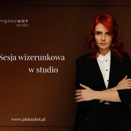Alicja Kopiec - Portret kobiety z rudymi włosami w czarnym garniturze, skrzyżowane ręce, studio z brązowym tłem, logo studia widoczne w lewym górnym rogu.