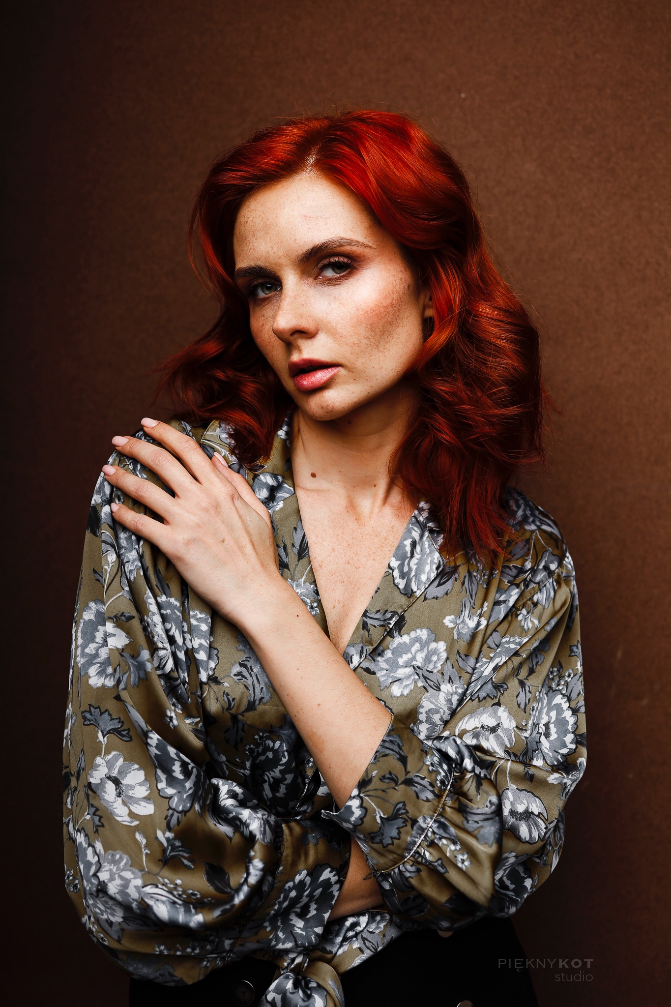 Portret kobiety z rudymi włosami i piegami, ubrana w kwiecistą koszulę na tle brązowej ściany, pozująca z ręką na ramieniu.