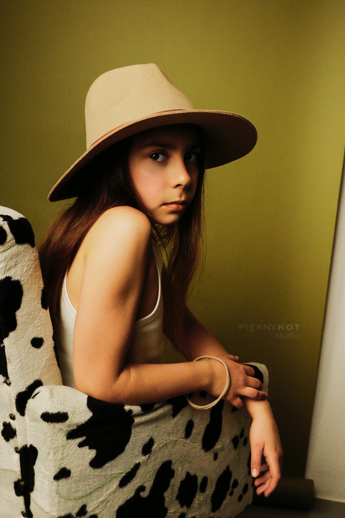 Portret dziewczynki w kapeluszu siedzącej na fotelu w łatki, z delikatnym makijażem i bransoletką, na oliwkowym tle.