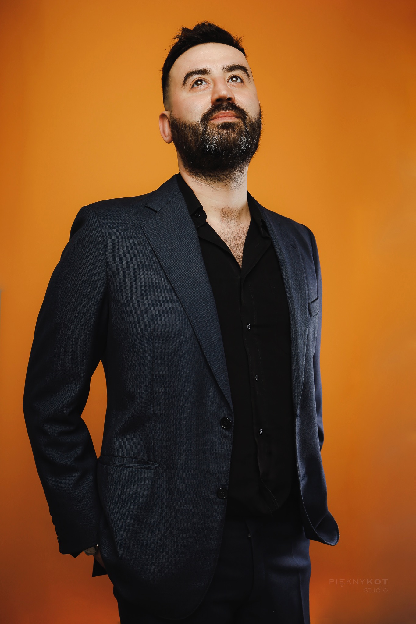 Mężczyzna z brodą w ciemnym garniturze i czarnej koszuli, patrzy w górę na pomarańczowym tle.