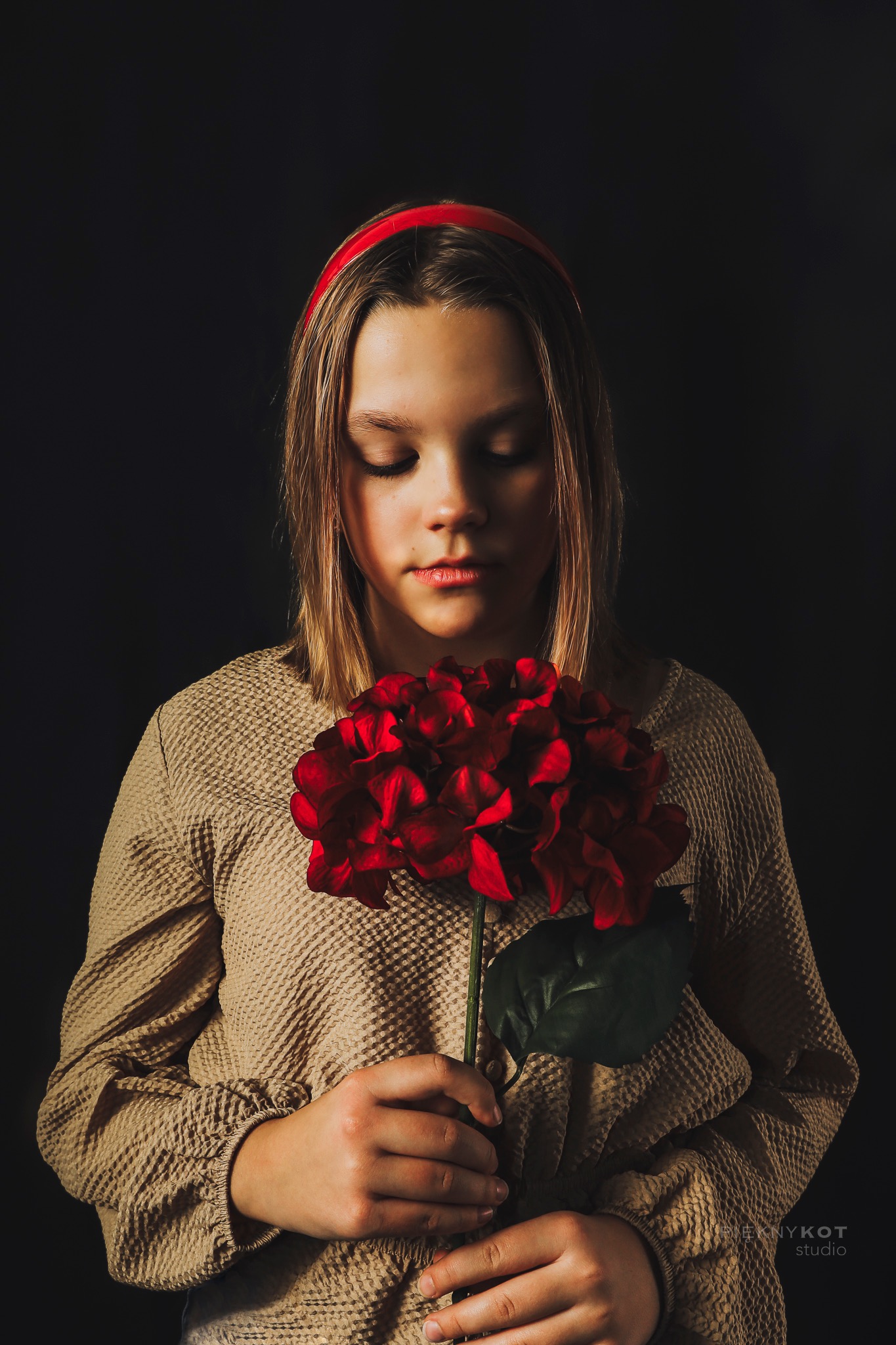 Portret młodej kobiety z zamkniętymi oczami, trzymającej czerwoną hortensję na ciemnym tle, z delikatnym oświetleniem podkreślającym rysy twarzy i fakturę sukienki.