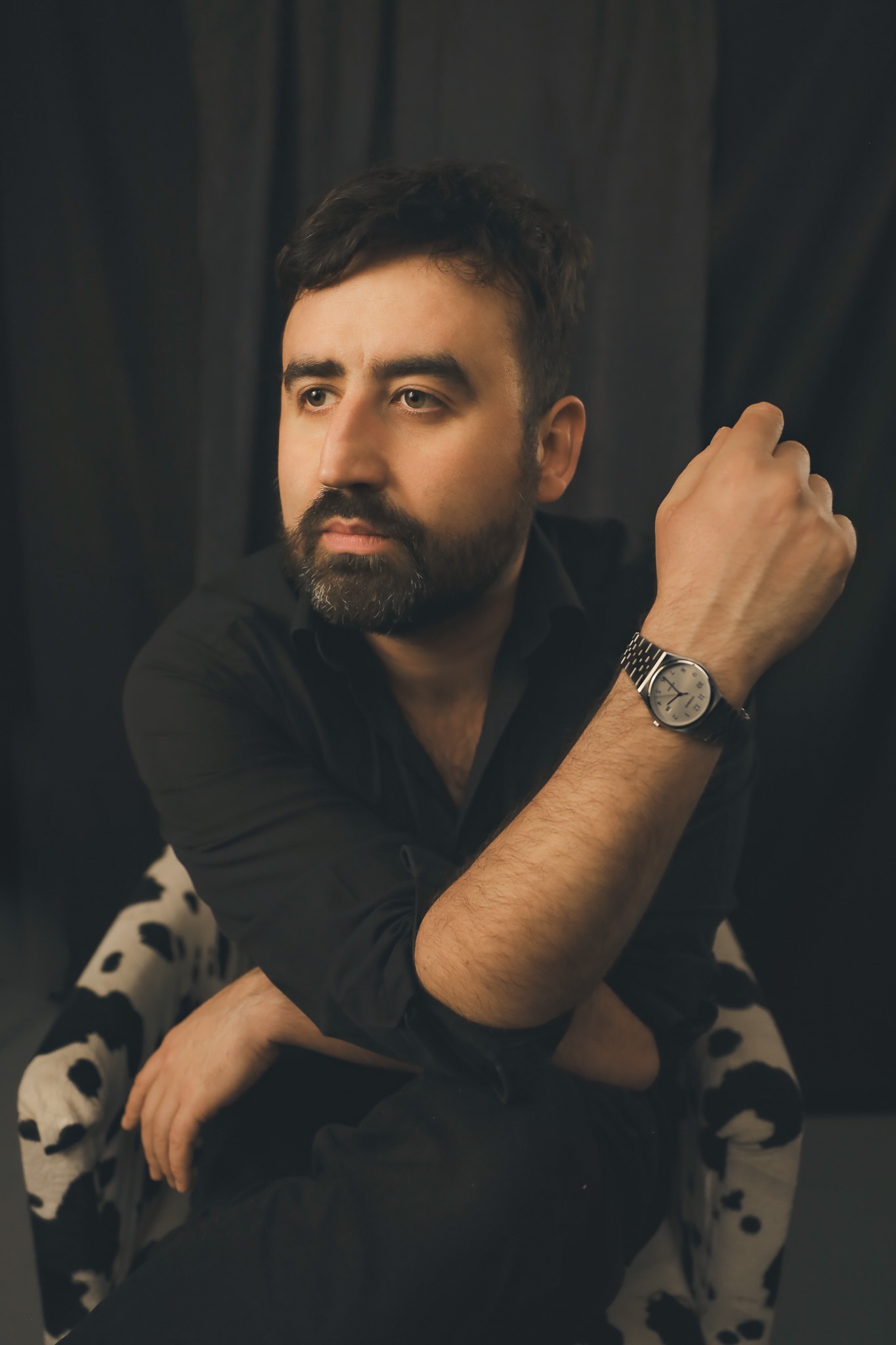 Portret mężczyzny w czarnej koszuli, siedzącego na krześle w czarno-białe łaty, z zegarkiem na ręce i zamyślonym spojrzeniem na tle ciemnego tła.