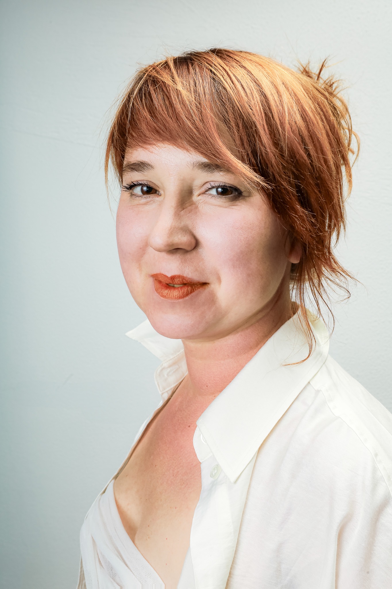 Portret kobiety z rudymi włosami, w białej koszuli, uśmiechająca się, na jasnym tle.