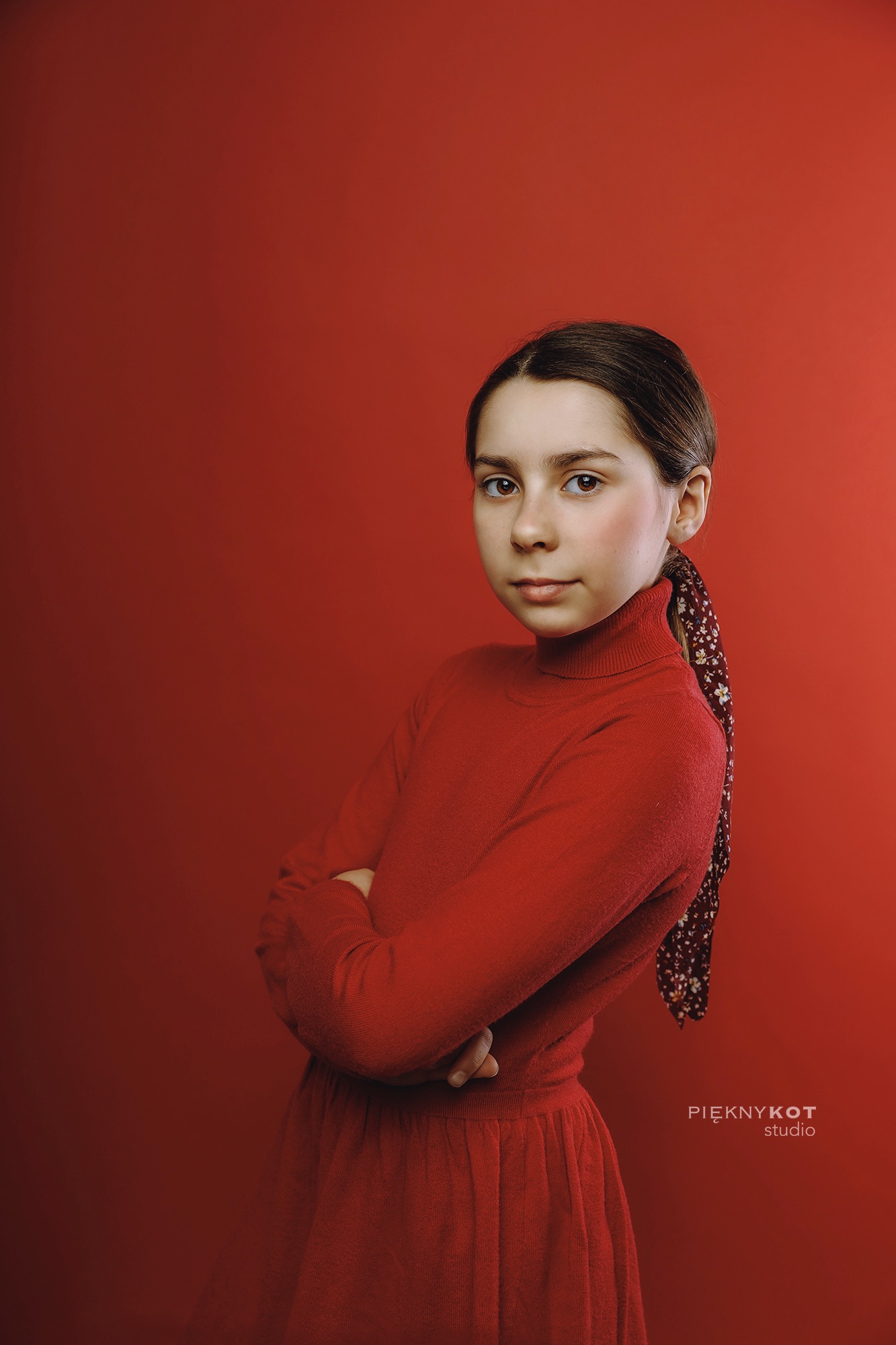 Portret młodej dziewczyny w czerwonym golfie i sukience, z chustą we włosach, na jednolitym czerwonym tle, pozująca z założonymi rękami.