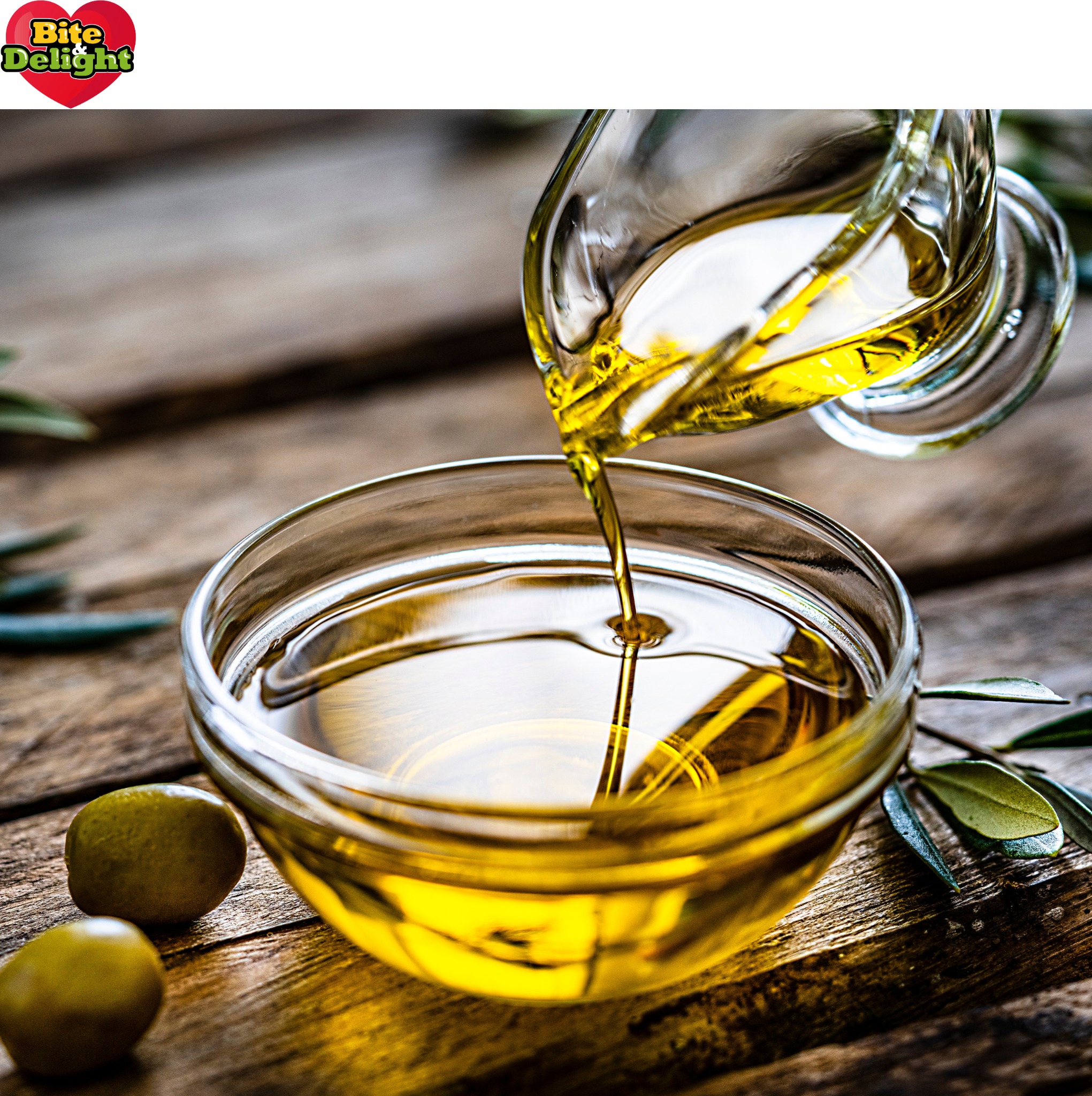 Strumień złocistej oliwy wlewany z przezroczystego dzbanka do szklanej misy, obok leżą dwie zielone oliwki i gałązka z liśćmi na rustykalnym, drewnianym stole.