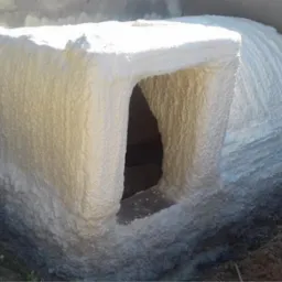 Fundament budynku z otworami okiennymi pokryty grubą warstwą białej pianki izolacyjnej, widoczne nierówności i struktura pianki, ziemia wokół fundamentu.