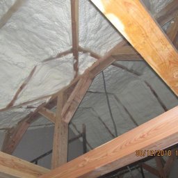 IZO PUR MAT Mateusz Oleksak - Poddasze z drewnianą konstrukcją pokryte białą, nierówną warstwą izolacji natryskowej, widoczne datowanie wykonania pracy w prawym dolnym rogu zdjęcia.