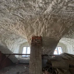 Poddasze po ociepleniu pianką PUR, widoczna nier&oacute;wnomierna struktura pianki na suficie i ścianach, ceglany komin na pierwszym planie, okna w tle.