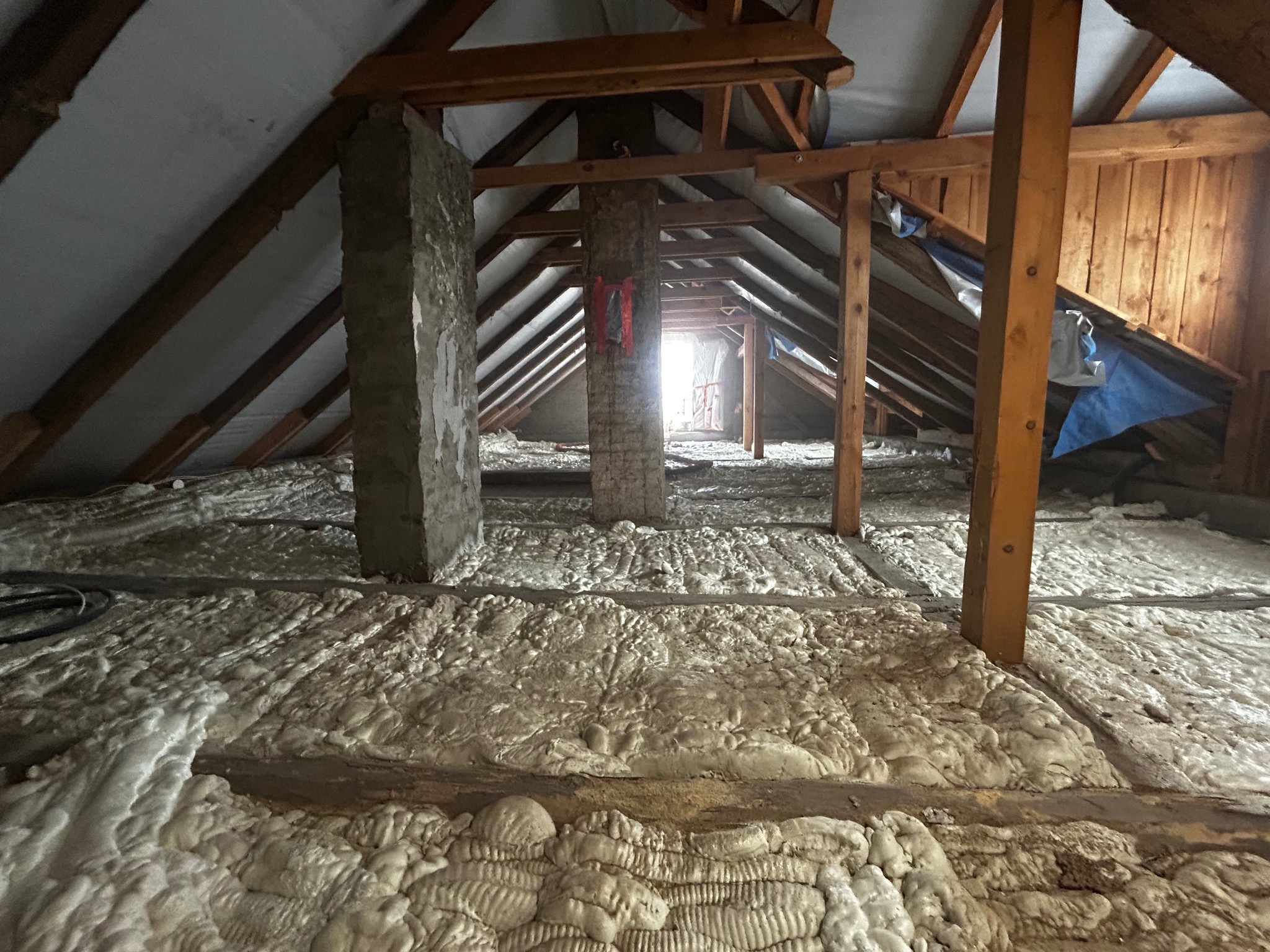 Poddasze z widoczną drewnianą konstrukcją dachu i kominem, pokryte grubą warstwą szarej piany izolacyjnej, z widocznymi elementami konstrukcyjnymi i fragmentami starej instalacji.