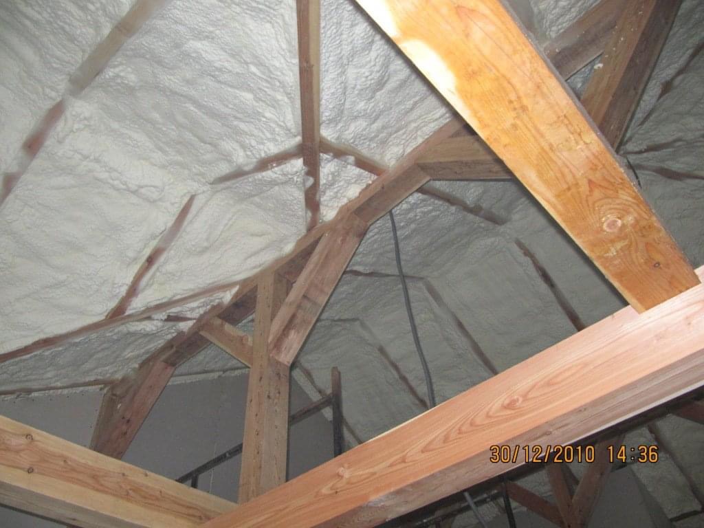 Poddasze z drewnianą konstrukcją pokryte białą, nierówną warstwą izolacji natryskowej, widoczne datowanie wykonania pracy w prawym dolnym rogu zdjęcia.