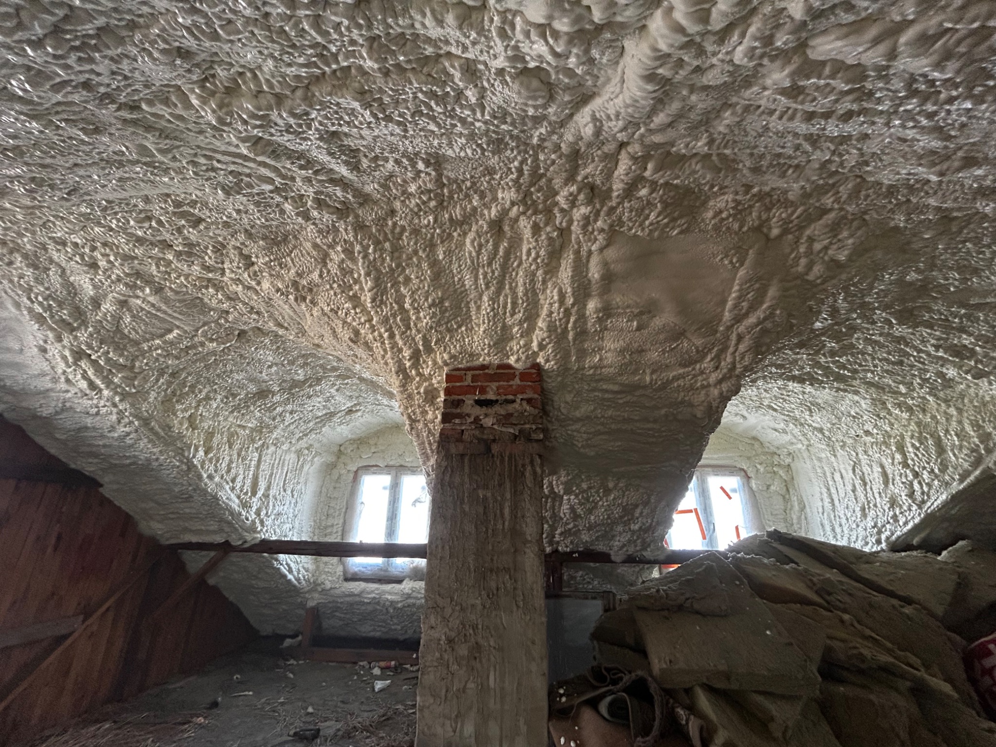 Poddasze po ociepleniu pianką PUR, widoczna nierównomierna struktura pianki na suficie i ścianach, ceglany komin na pierwszym planie, okna w tle.