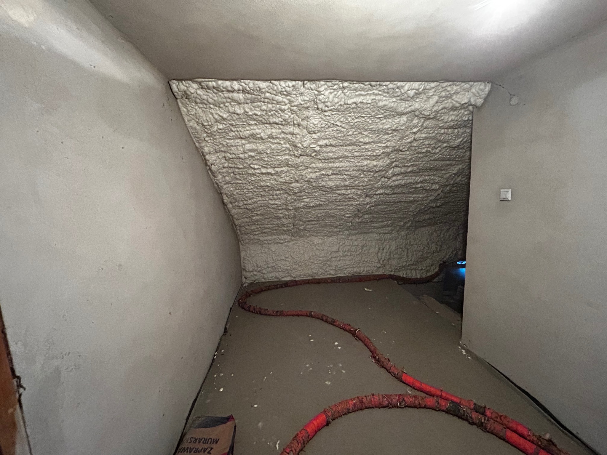 Poddasze po natrysku pianki izolacyjnej, widoczne nierówności i struktura piany, czerwony wąż na podłodze, worek zaprawy murarskiej w rogu.