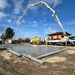 Wylewanie fundament&oacute;w pod budynek z użyciem pompy do betonu, widoczne zbrojenia i praca ekipy budowlanej w słoneczny dzień.