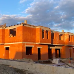 Surowy stan dwukondygnacyjnego budynku z czerwonej cegły ceramicznej w trakcie budowy, z widocznymi otworami okiennymi i drzwiowymi, na tle błękitnego nieba z obłokami.