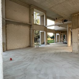 Bartłomiej Melon - Wnętrze surowego stanu budynku z betonu komórkowego i betonu, widoczne surowe ściany z pustaków, betonowe słupy i strop, drabina oparta o ścianę, na zewnątrz zieleń.