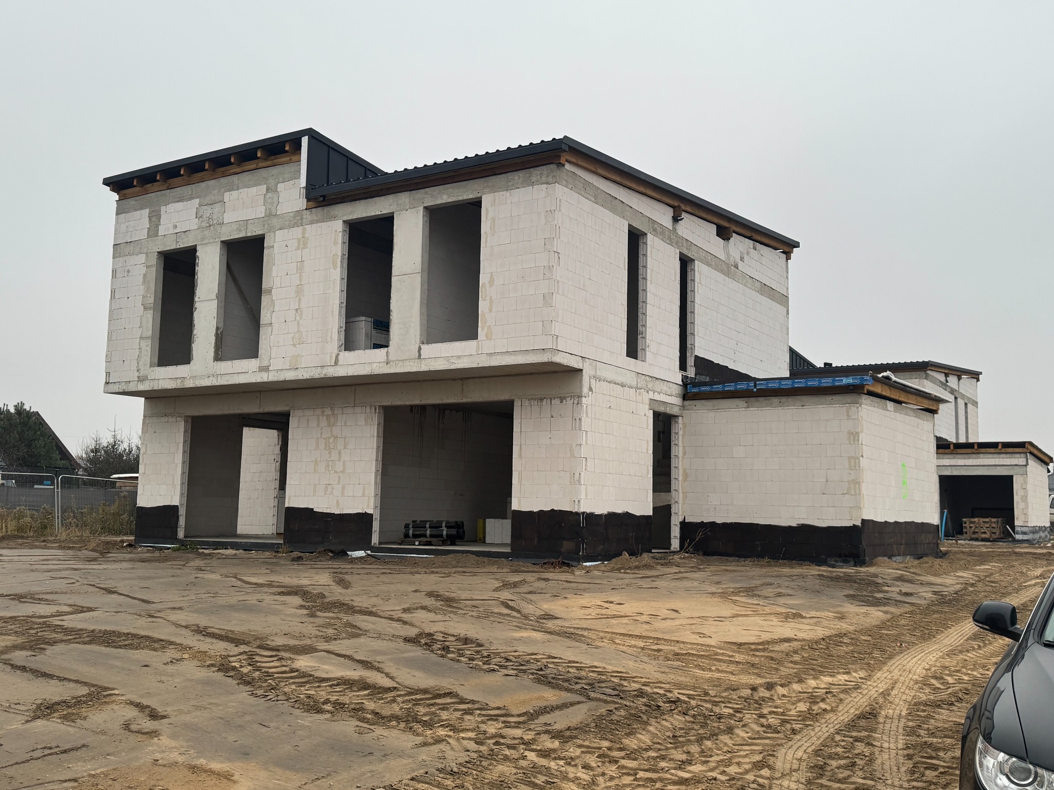 Surowy stan dwukondygnacyjnego budynku z białych bloczków, widoczne otwory okienne i drzwiowe, czarna hydroizolacja fundamentów, teren budowy z widocznymi śladami opon.