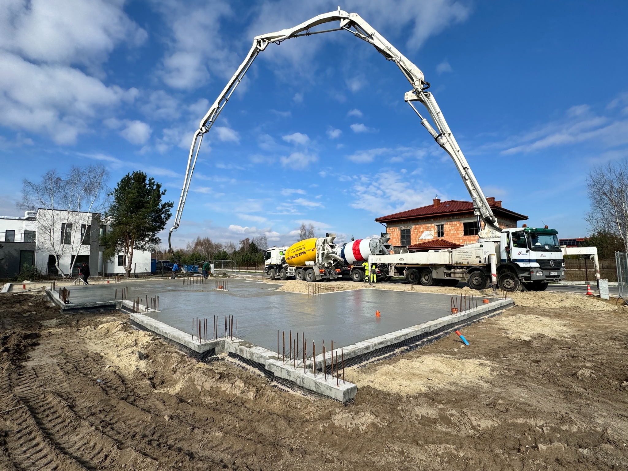 Wylewanie fundamentów pod budynek z użyciem pompy do betonu, widoczne zbrojenia i praca ekipy budowlanej w słoneczny dzień.