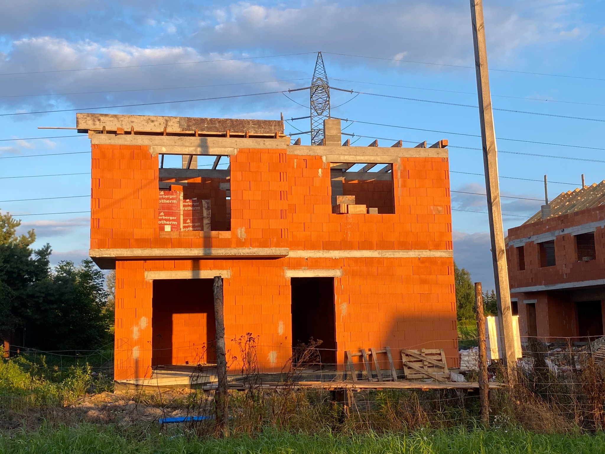 Surowy, dwukondygnacyjny budynek z cegły ceramicznej w trakcie budowy, z widocznymi paletami cegieł na górnej kondygnacji, z słupem elektrycznym i liniami wysokiego napięcia w tle.